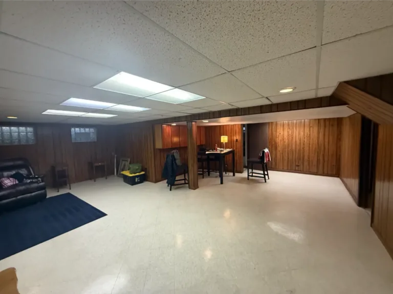 Open floor area of before image of basement showing a recliner in one corner, with a table and a lamp with a couple chairs around it in the far corner center.