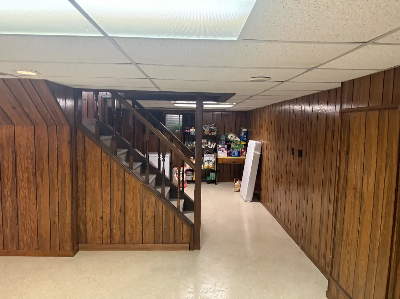 Before image of basement area nearby the stairs, with a small mattress and kitchen odds and ends including paper towels and snacks on tables in the background.