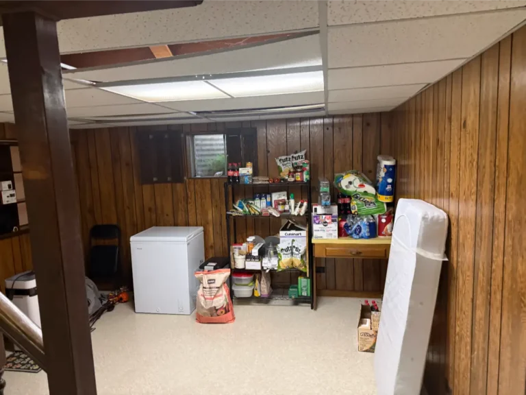 Before image of a room within a basement that has tile flooring, an ice chest, and various kitchen odds and ends like snacks, paper towels, bottles of water and so on.