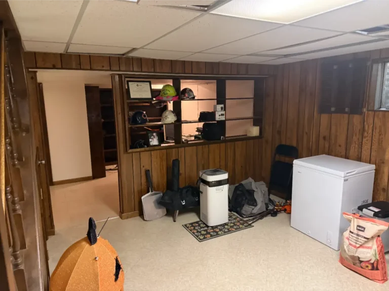 Before image of a basement with tile floors, an ice chest and construction style hats on a series of shelves in the background. Various items lay about the floor.